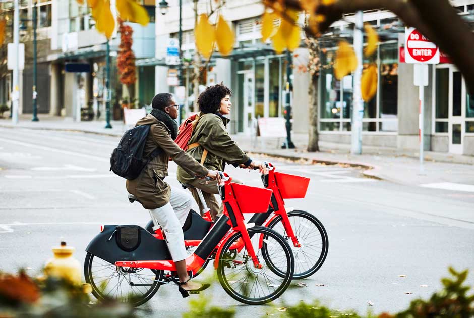 A pair of women glide forward on bikes in a busy shopping district.